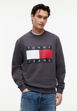 Joven vestido con sudadera gris oscuro con gran logo de Tommy Jeans y vaqueros azul claro, posando frente a un fondo blanco.