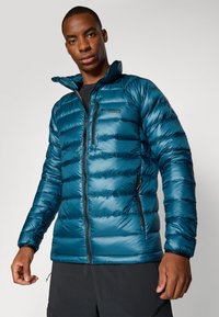 Teal pufferjakke med vandret syninger, lynlåslomme på brystet og sidelommer. Har en stående krave og sorte accenter.