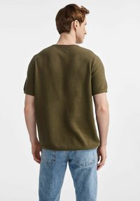 Oliwkowa dzianinowa koszulka o luźnym kroju, z krótkimi rękawami i okrągłym dekoltem. Noszona z jasnoniebieskimi dżinsami.