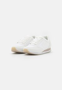 Weiße Sport Schuhe mit einem glatten synthetischen Obermaterial, Schnürsenkeln, einer strukturierten Gummisohle und einem beigen sowie grauen Akzentstreifen entlang der Sohle.