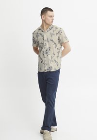 Camisa de manga corta beige con estampado floral y acentos en azul y negro, combinada con pantalones azul marino y zapatos de ante grises, modelo de pie de lado.