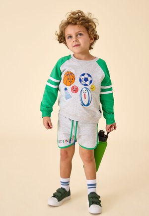 Fiatal gyermek göndör hajjal, szürke és zöld sportöltözetben, rajzfilm-labdákkal díszítve, zöld vizes palackot tartva bézs háttér előtt.