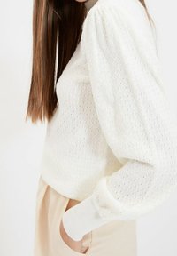 Pull blanc texturé avec motifs en losange, manches bouffantes et poignets côtelés. Associé à un pantalon clair, ajusté.