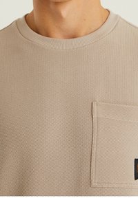 Beige strukturovaná mikina s žebrovaným kulatým výstřihem, náprsní kapsou na levé straně a malým logem v dolním pravém rohu.