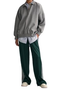 Graues Sweatshirt mit Polo-Kragen und drei Knöpfen, über einem hellblauen Hemd getragen. Kombiniert mit grünen Sporthosen mit seitlichen Streifen.