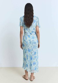 Blumenkleid in Hellblau mit blauen Rosen, kurzen Ärmeln, einem figurbetonten Oberteil und einem ausgestellt geschnittenen Saum. Das Model trägt beige Sandalen.