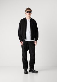 Schwarze Bomberjacke mit Reißverschluss, darunter ein weißes T-Shirt, schwarze Cargo-Hosen und schwarze klobige Schuhe. Model trägt schwarze Sonnenbrille.