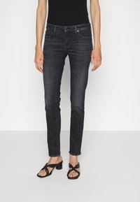 Jeans de mezclilla negra de corte ajustado, con un cierre de botón clásico, cinco bolsillos y un ligero desgaste. Combinados con sandalias de tacón negras.