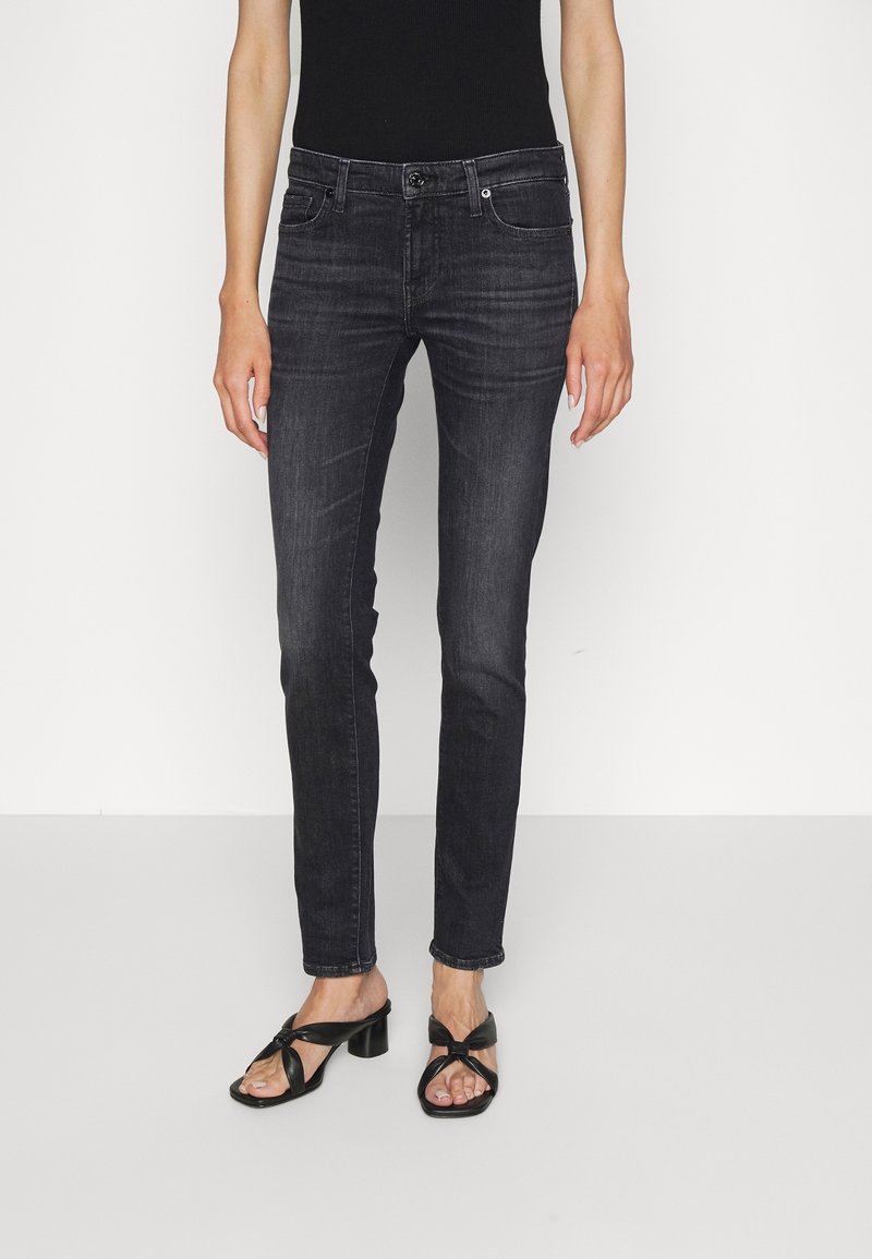 Jeans de mezclilla negra de corte ajustado, con un cierre de botón clásico, cinco bolsillos y un ligero desgaste. Combinados con sandalias de tacón negras.