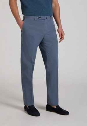 Uomo con pantaloni blu aderenti con passanti per cintura e mocassini blu scuro, in piedi con una mano in tasca.