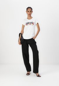 T-shirt blanc avec un graphisme floral et le texte "LIU JO", associé à un pantalon noir à jambe large. Les accessoires comprennent un sac à main noir et des sandales à talons.
