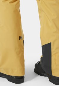 Gele waterdichte skibroek met zwarte accenten, voorzien van een gladde textuur, Helly Tech merknaam en beschermende enkelbedekking over zwarte laarzen.