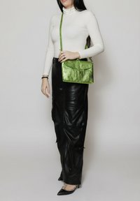 Borsetta metallica verde con chiusura a soffietto e tracolla regolabile. Indossata con un maglione aderente bianco a collo alto e pantaloni in pelle nera. Calzature a punta.