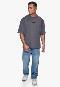 Mørkegrå oversized T-shirt med korte ærmer, der har et lille sort logo-patch på brystet. Kombinationen med løse blå denimjeans og sneakers.