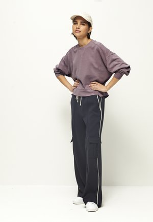 Young person wearing a light beige cap, oversized mauve sweatshirt, black wide-leg pants with white side stripes, posing against a white background.