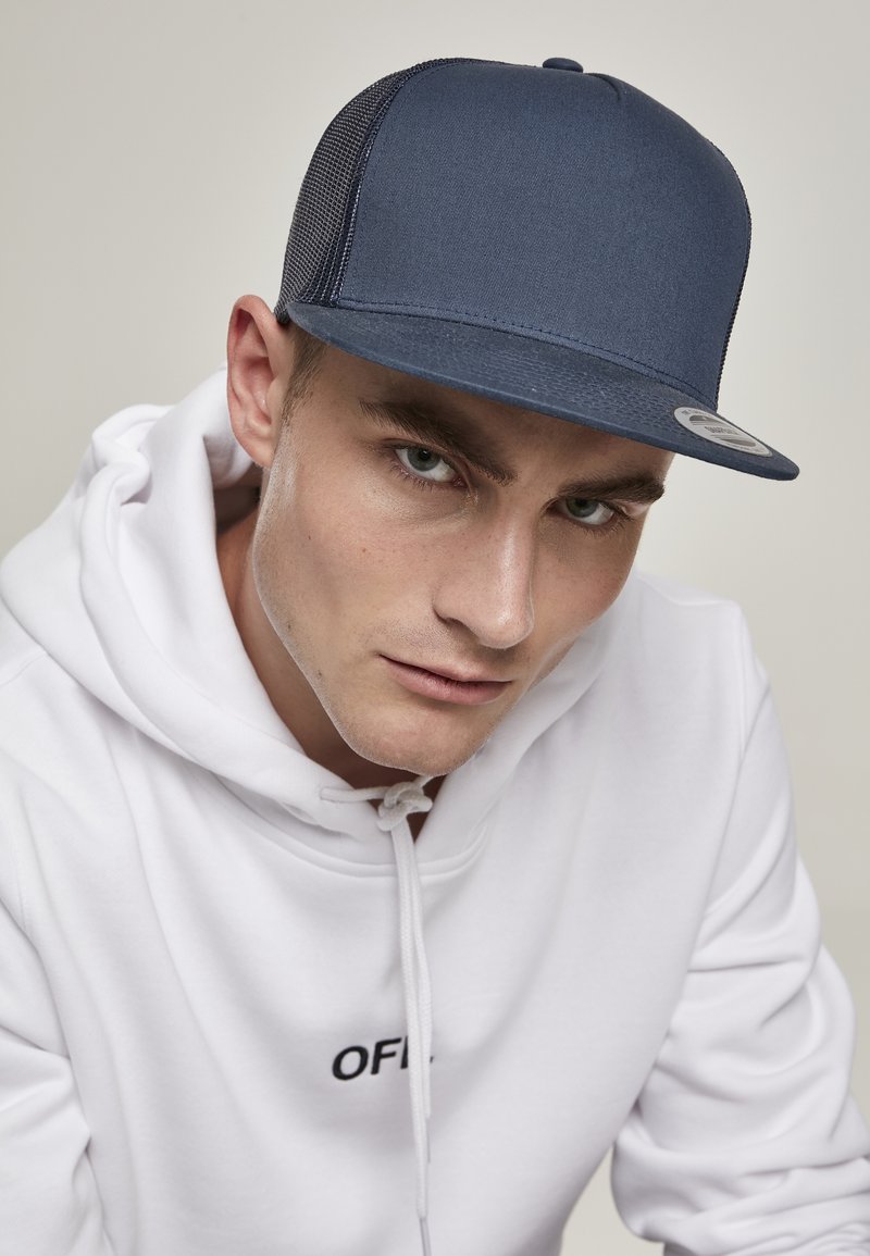 Casquette bleu marine à l'arrière en maille avec une visière plate, tissu texturé, couture contrastante et un logo circulaire en patch sur le côté. Portée avec un sweat-shirt blanc.
