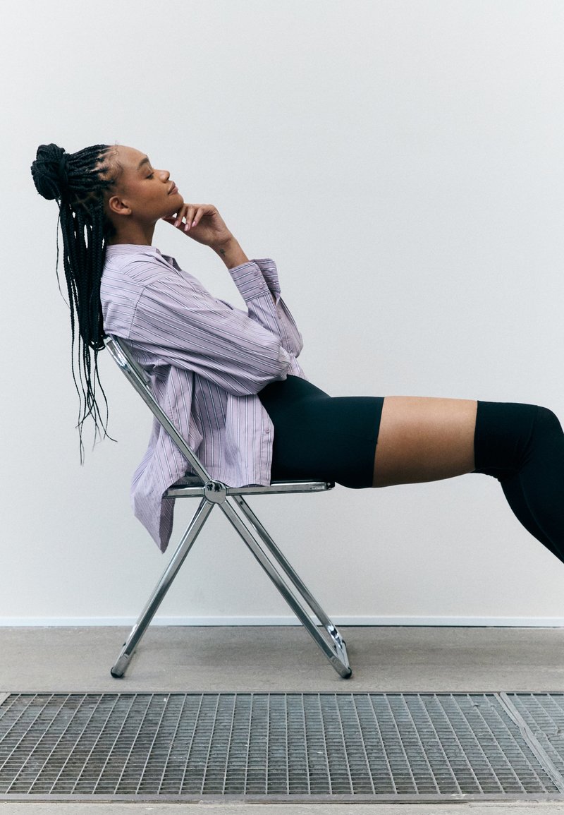 Una silla plegable con patas de cromo sostiene a una persona con una camisa de rayas moradas sueltas y leggins negros, con el cabello largo trenzado que cae por su espalda.