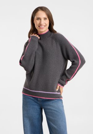 Smiling woman with brown hair wears a gray knit sweater with pink and purple stripes and blue jeans, posing with one hand on her hip.
