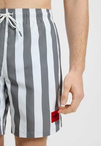 Zwemshorts in grijs en witte verticale strepen met een elastische tailleband, koord en een rode "HUGO" logo patch aan de zijkant.
