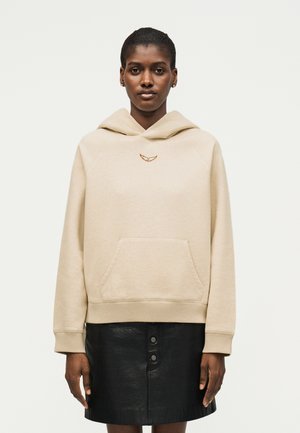 Γυναίκα που φοράει μπεζ φούτερ με κουκούλα με μικρό κεντημένο λογότυπο και μαύρη δερμάτινη φούστα με κουμπιά, στέκεται μπροστά σε απλό ανοιχτό φόντο.