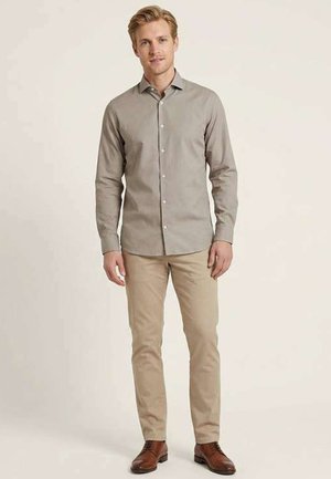 Homme debout portant une chemise beige clair à boutons, un pantalon kaki et des chaussures habillées marron sur un fond uni.