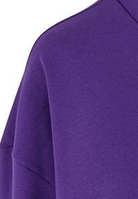 Gros plan sur le tissu d'un sweat-shirt à col ras du cou violet, montrant la couture de l'épaule et la texture côtelée du col.
