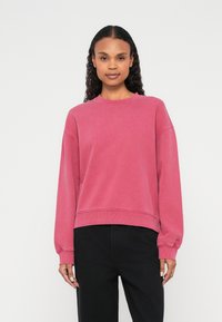 Różowa bluza z długimi rękawami, prążkowanym dekoltem i mankietami, casualowy krótki fason. W zestawie z czarnymi spodniami o luźnym kroju.