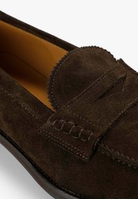 Mocasines de ante marrón con punta redondeada, costuras tonales y una correa decorativa. El forro interior es de un amarillo contrastante. Textura suave.