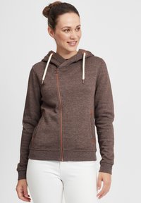 Brun zip-up hettegensar laget av mykt stoff. Har hette, kremfargede snorer og brune lærdetaljer. Figurtilpasset design med sidelommer.
