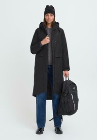 Svart lång kappa med huva och framfickor, ihop med en grå tröja, blå jeans, svarta kängor och en svart ryggsäck. Enkelt design.