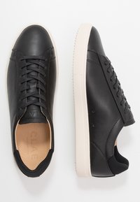 Black leather sneakers with a smooth finish, featuring a beige rubber sole, contrasting stitching, and a rounded toe. Laces and branding visible.