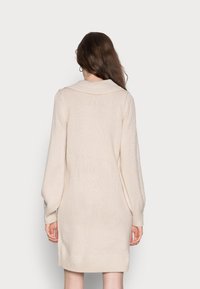 Abito lavorato a maglia beige chiaro con un colletto morbido e sovradimensionato e maniche lunghe leggermente a sbuffo. Orlo e polsini a coste, tessuto testurizzato.