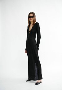 Schwarzes Kleid mit langen Ärmeln, tiefem V-Ausschnitt und seitlichen Schlitzen, strukturierter Stoff, figurbetonte Silhouette, kombiniert mit spitzen schwarzen Ballerinas.