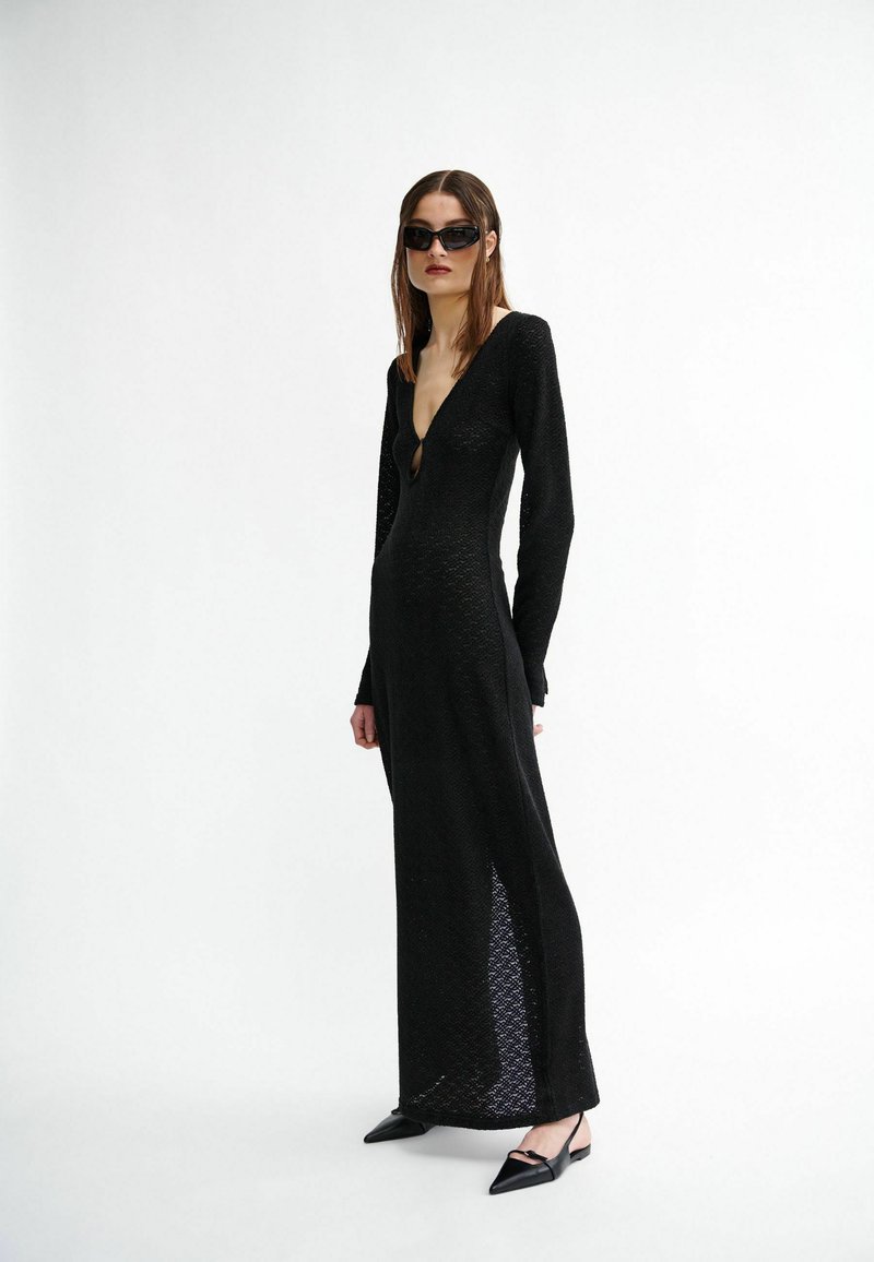 Schwarzes Kleid mit langen Ärmeln, tiefem V-Ausschnitt und seitlichen Schlitzen, strukturierter Stoff, figurbetonte Silhouette, kombiniert mit spitzen schwarzen Ballerinas.