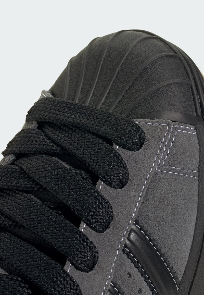 Svart mockasinsneaker med texturerad svart gummitätning vid tån, svarta snören och kontrasterande vit söm längs sidan.