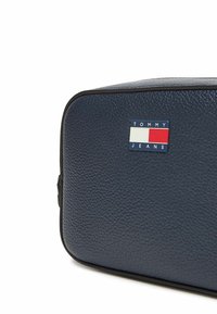 Borsa da toilette in pelle blu navy con superficie testurizzata, bordi neri e forma rettangolare. Presenta un logo Tommy Jeans blu, bianco e rosso.