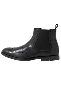 Zwarte leren Chelsea-boot met elastische zijpanelen, decoratieve brogue-details, ronde neus en een lage gestapelde hak. Gladde textuur overal.