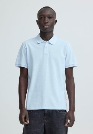 Hombre vestido con una camisa polo de manga corta azul claro y pantalones oscuros, de pie contra un fondo liso gris claro, mirando hacia adelante.