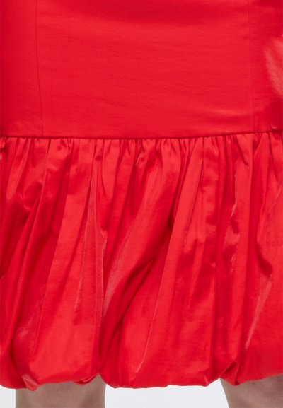 Jupe à ourlet boule rouge avec une texture de tissu brillante et froncée, couvrant partiellement les jambes visibles en bas.