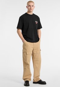 T-shirt nera in cotone con piccolo logo sul petto, abbinata a pantaloni cargo beige con molteplici tasche. Il modello indossa scarpe nere.