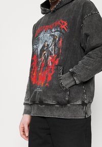 Pullover in felpa grigia scura in cotone misto, con una grafica frontale raffigurante una figura scheletrica, accentuata da fiamme rosse e una tasca a marsupio.