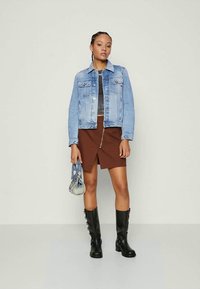 Young woman wearing a light blue denim jacket, brown skirt with front slit, black boots, holding a small blue handbag, standing against plain background.