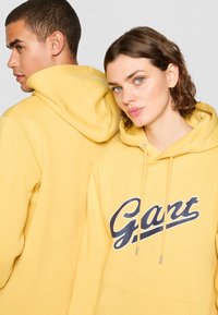 Gule hettegensere med frontlomme. Har en marineblå logo "Gant" over brystet. Mykt bomullsmateriale, avslappet passform.