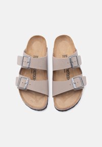 Μπεζ σανδάλια Birkenstock με διπλό λουράκι, ρυθμιζόμενες ασημένιες πόρπες και ανάγλυφα φελλοποδία, εμφανιζόμενα από επάνω σε λευκό φόντο.