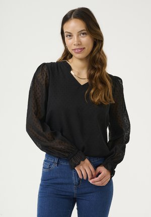 Black blouse with textured polka dot pattern, V-neckline, and sheer puffed sleeves, paired with blue jeans. Subtle elastic cuffs.