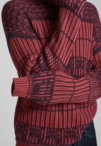 Gestrickter Pullover mit einem geometrischen Muster in Burgunderrot und Dunkelblau, mit hohem Kragen und gerippten Bündchen für Struktur und Wärme.