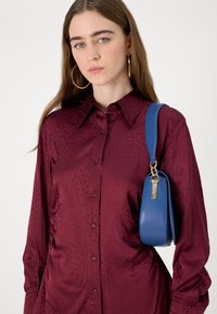 Camisa bordô com padrão discreto, mangas longas e design abotoado. Bolsa azul com textura suave e detalhe em metal dourado.