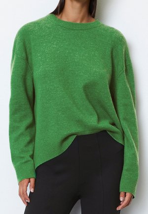 Mujer que lleva un suéter de punto de manga larga de color verde brillante metido dentro de unos pantalones negros de cintura alta sobre un fondo liso.