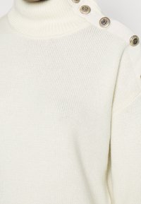 Crèmmönstrad turtlenecktröja med silverfärgade dekorativa knappar längs axelsömmen.