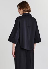 Una donna con capelli corti castani che indossa una camicia nera larga con maniche ampia e pantaloni neri, in piedi contro uno sfondo chiaro e semplice.